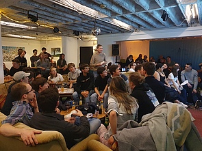 Foto vom SciencePub Quiz. Es sind viele glückliche Menschen im Innenraum eines Cafes zu sehen. Sie schauen auf Vincent, einen großen Mann, der lachend in der Mitte des Raums steht und moderiert.