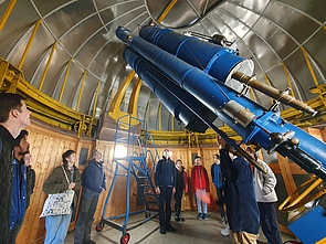 Innenraum einer Kuppel mit großem blauem Teleskop. Eine Gruppe junger Menschen steht am Rand des Raums und schaut auf das Teleskop.